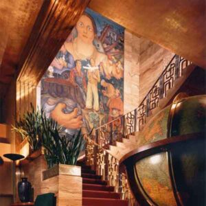 Staircase with a large mural painted on the back wall. Mural shows a woman's head and bare shoulders at the top, holding fruit in her left hand. Smaller figures of men are shown below, engaged in different activities. One man has a small airplane, and some are working in fields.
