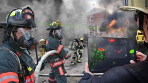 A house on fire, with flames and black smoke coming out of it. Four firefighters are holding hoses in front of the house. A fifth firefighter is holding a tablet showing green outlines of firefighters inside the house, one walking up the stairs. The tablet says “MAYDAY” at the top and shows various controls, maps, and graphs.
