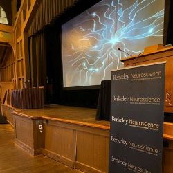 A stage with an artistic rendering of a neuron on the screen, and a sign in front of the podium that says "Berkeley Neuroscience" and "Helen Wills Neuroscience Institute" repeating on it.
