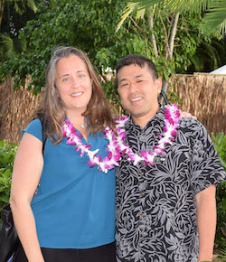 Tracy and Kauwe are wearing leis and standing outside in front of greenery.