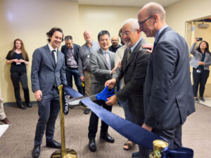 Feinberg smiling, cutting a ribbon with giant scissors, surrounded by other smiling people.