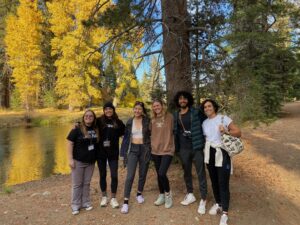 6 smiling students standing on a trail by water, with fall foliage in the background.